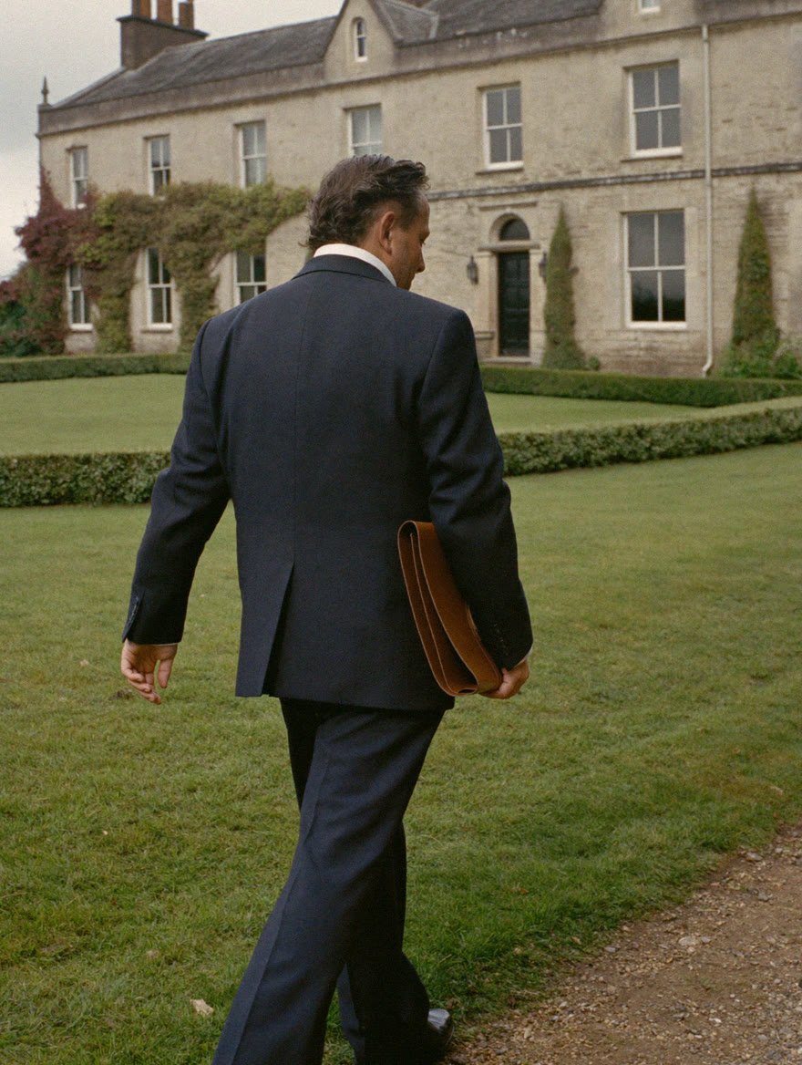 An estate manager in a navy suit walking the lawn toward a stone country house